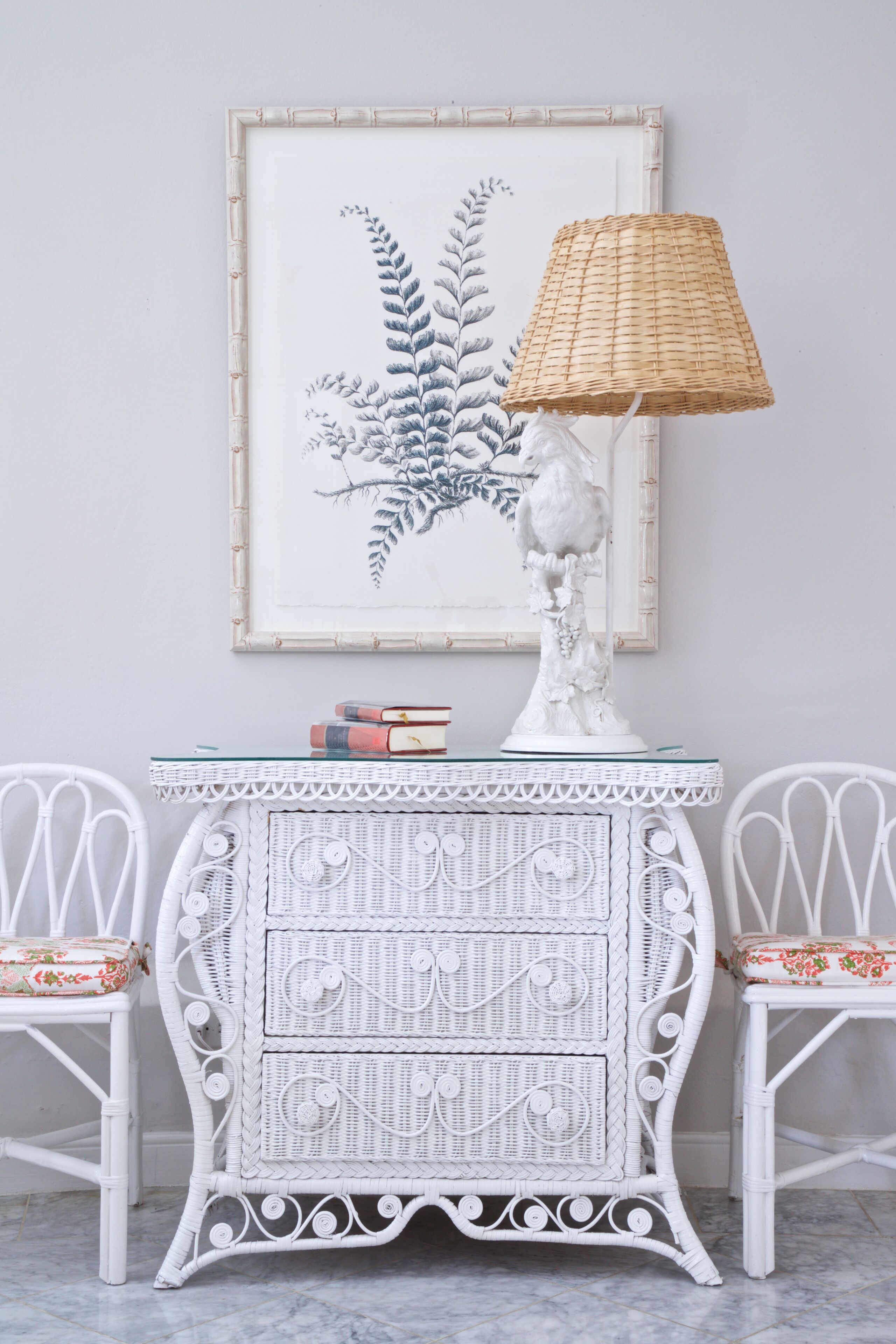 Una mesita auxiliar de mimbre blanco con una lámpara y libros, flanqueada por dos sillones de mimbre blanco con cojines estampados.