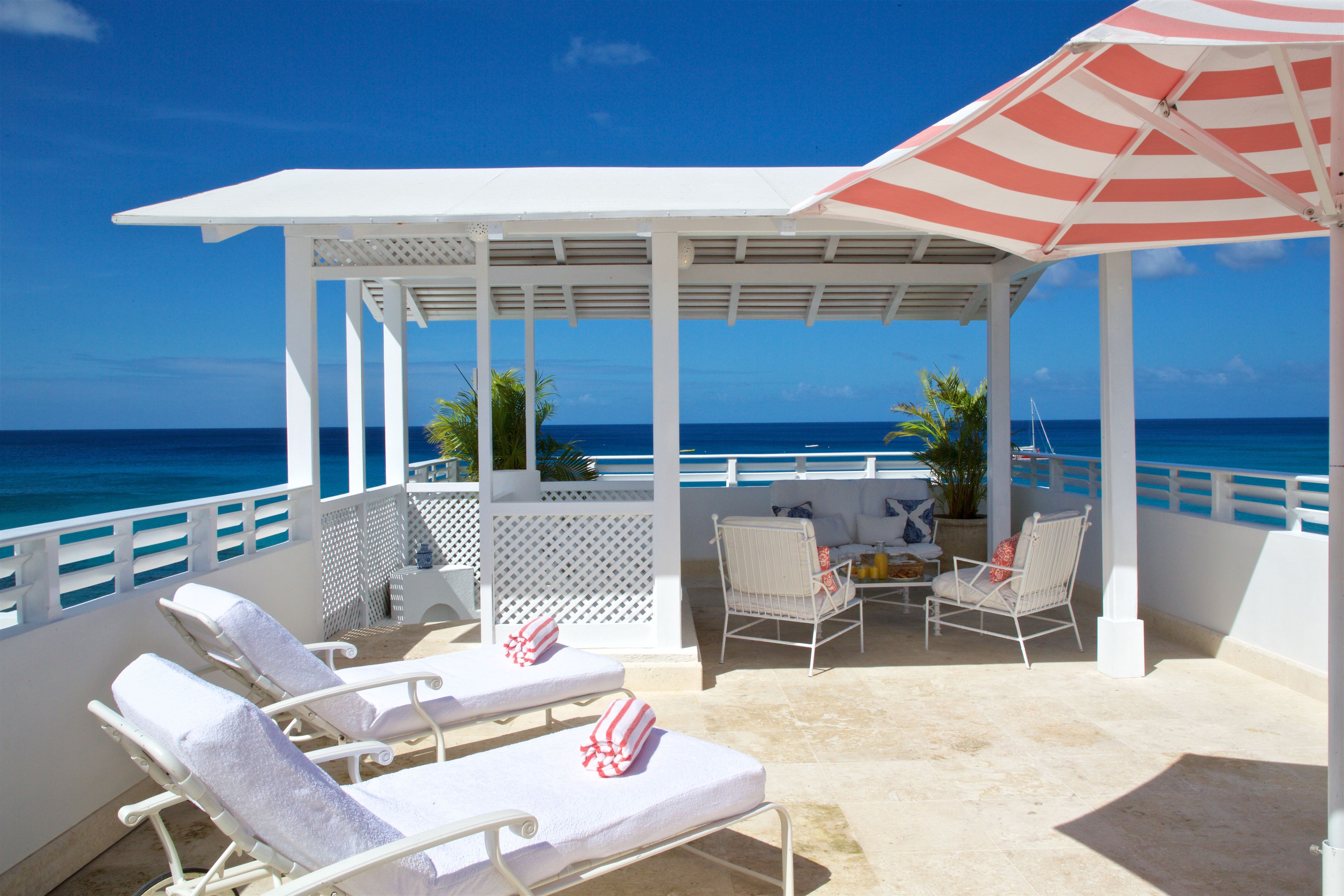 A covered outdoor seating area with lounge chairs and a patio umbrella, overlooking the ocean.