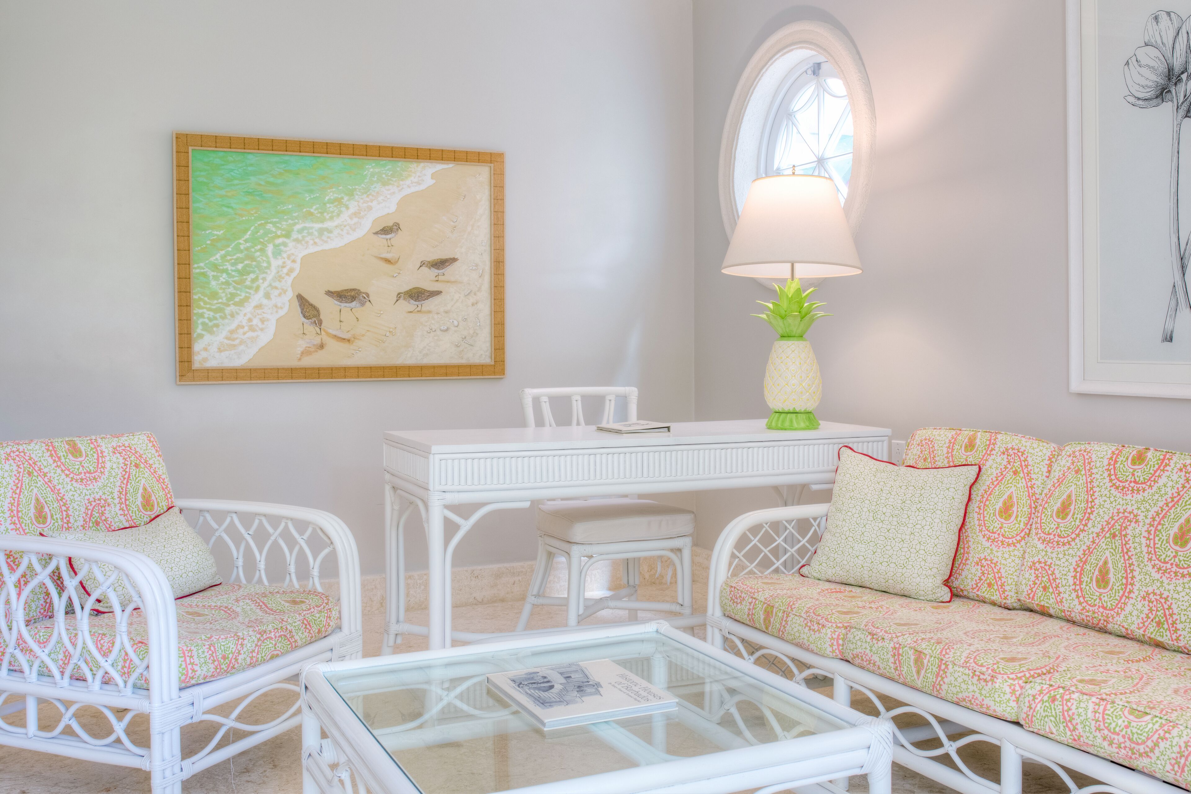 Um quarto com um sofá e uma cadeira de vime branco, uma mesa de centro de vidro, uma escrivaninha branca e um quadro de uma praia.