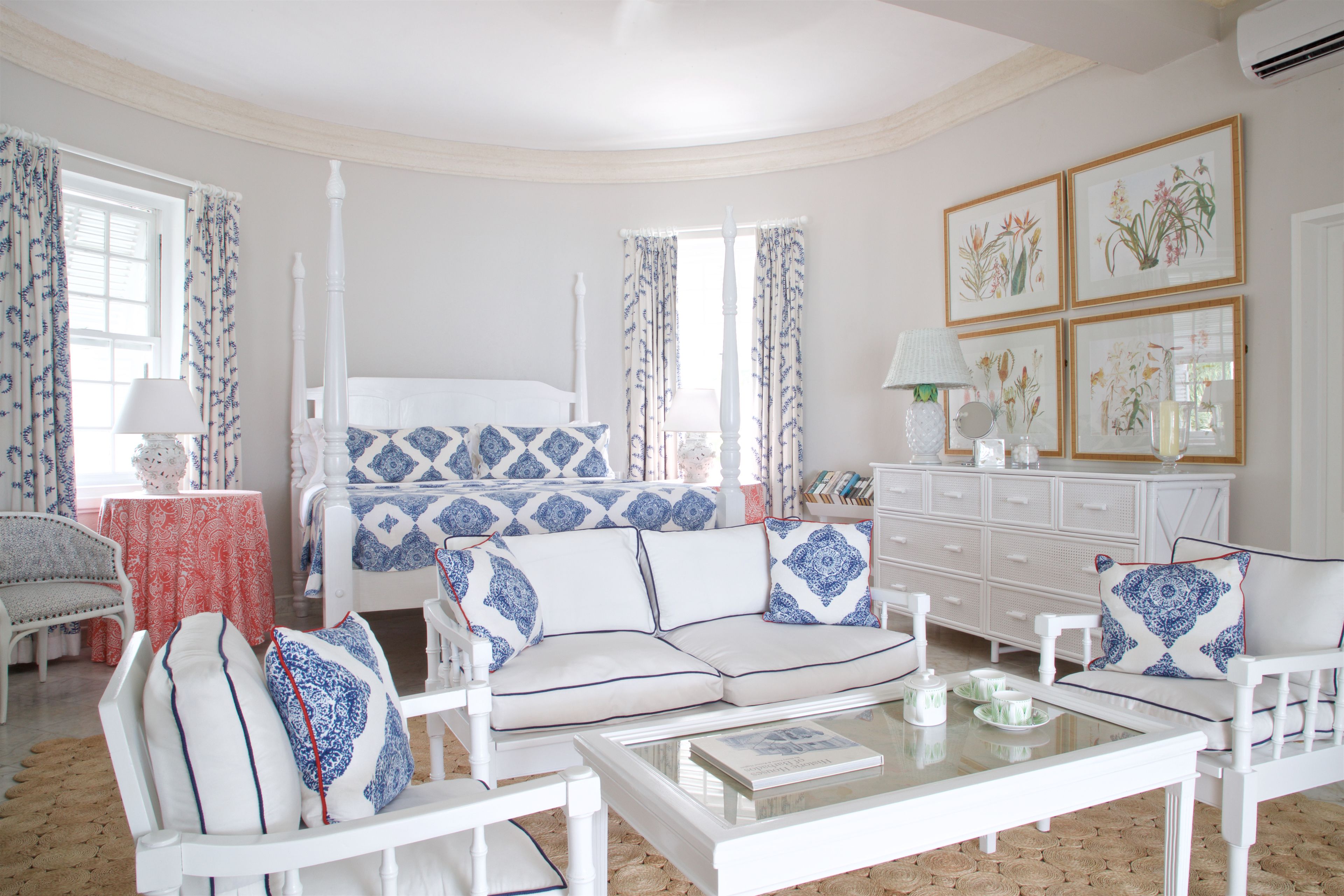 Una habitación con una cama con dosel blanco, un sofá blanco con cojines estampados en azul y blanco, un sillón blanco, una mesa de centro de cristal y un aparador blanco con espejo.
