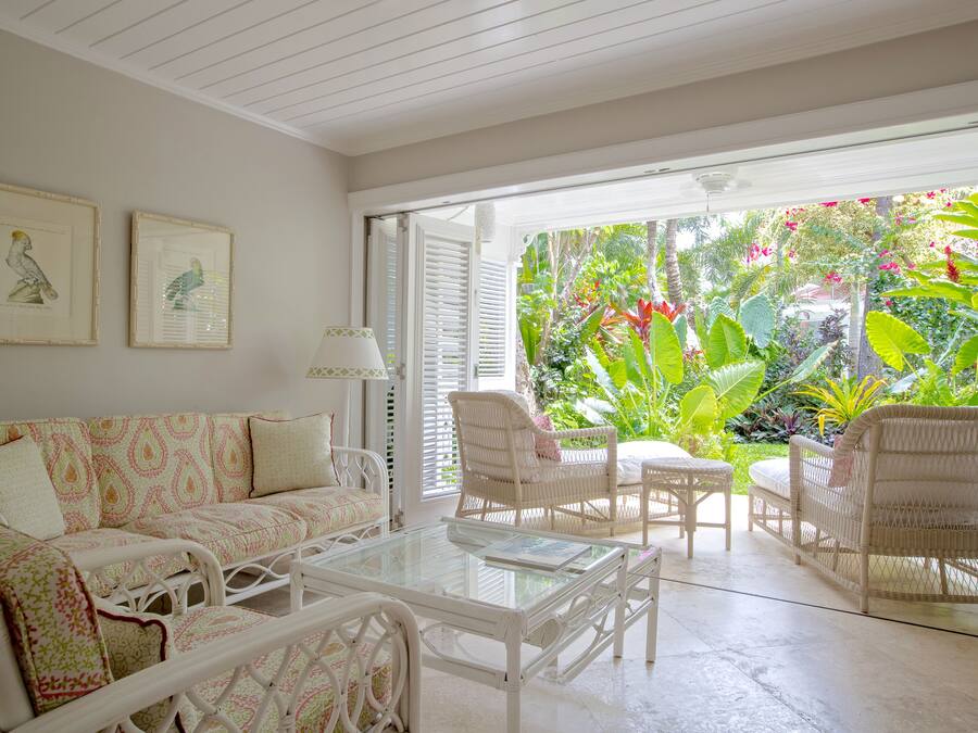 Een ruime woonkamer met een witte rieten bank, een glazen salontafel en rieten stoelen. Twee kunstwerken hangen in lijsten aan de muur, en de kamer grenst aan een weelderige tuin.
