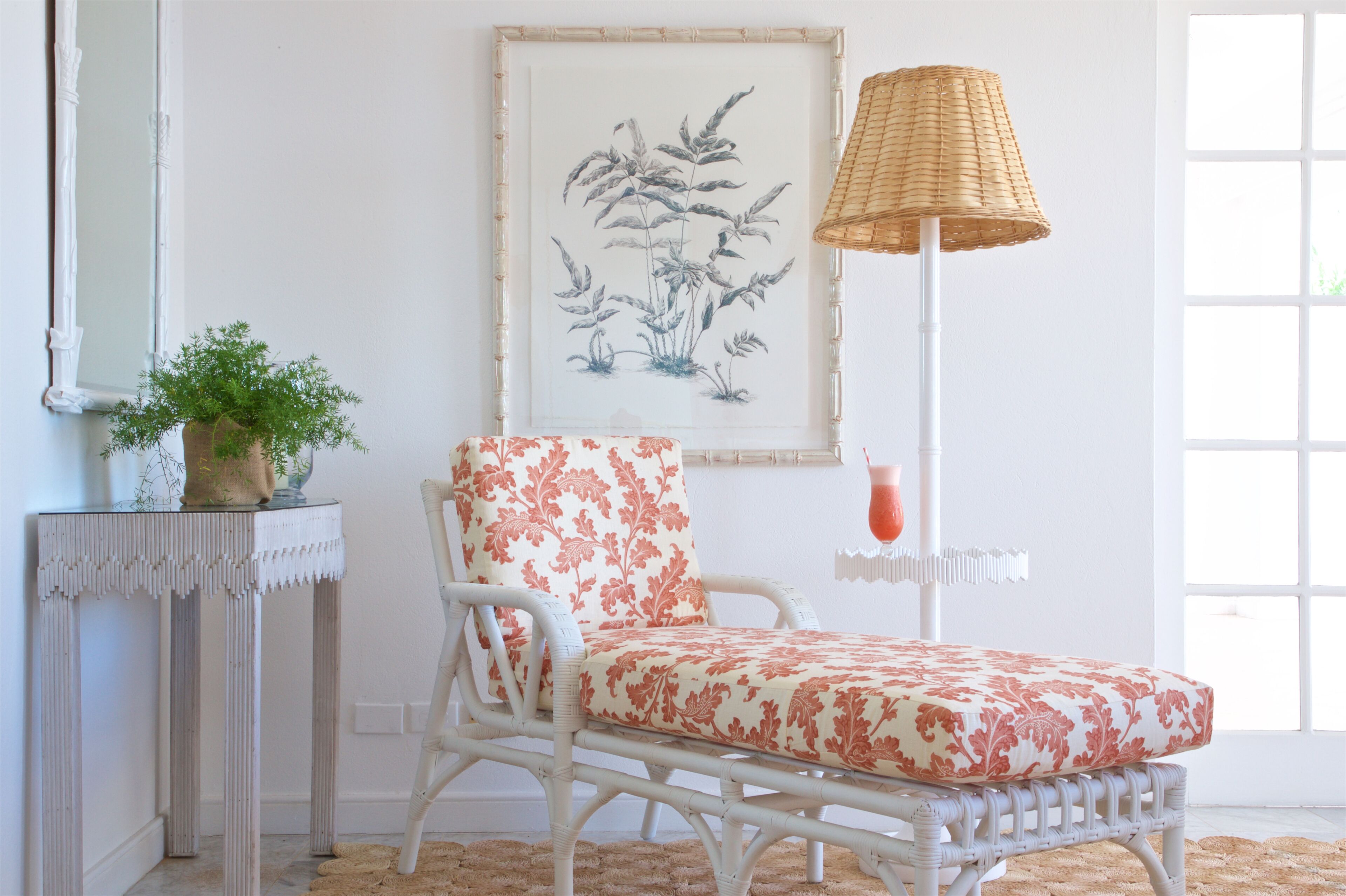 Una silla de mimbre blanco con un cojín floral, una planta en maceta sobre una consola blanca y una impresión botánica enmarcada en la pared.