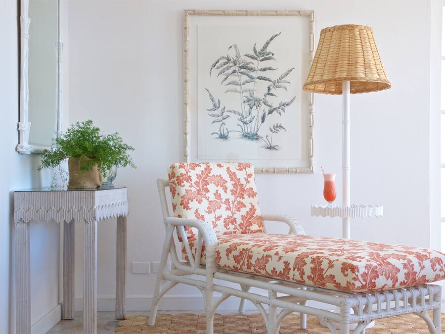 Una silla de mimbre blanco con un cojín floral, una planta en maceta sobre una consola blanca y una impresión botánica enmarcada en la pared.