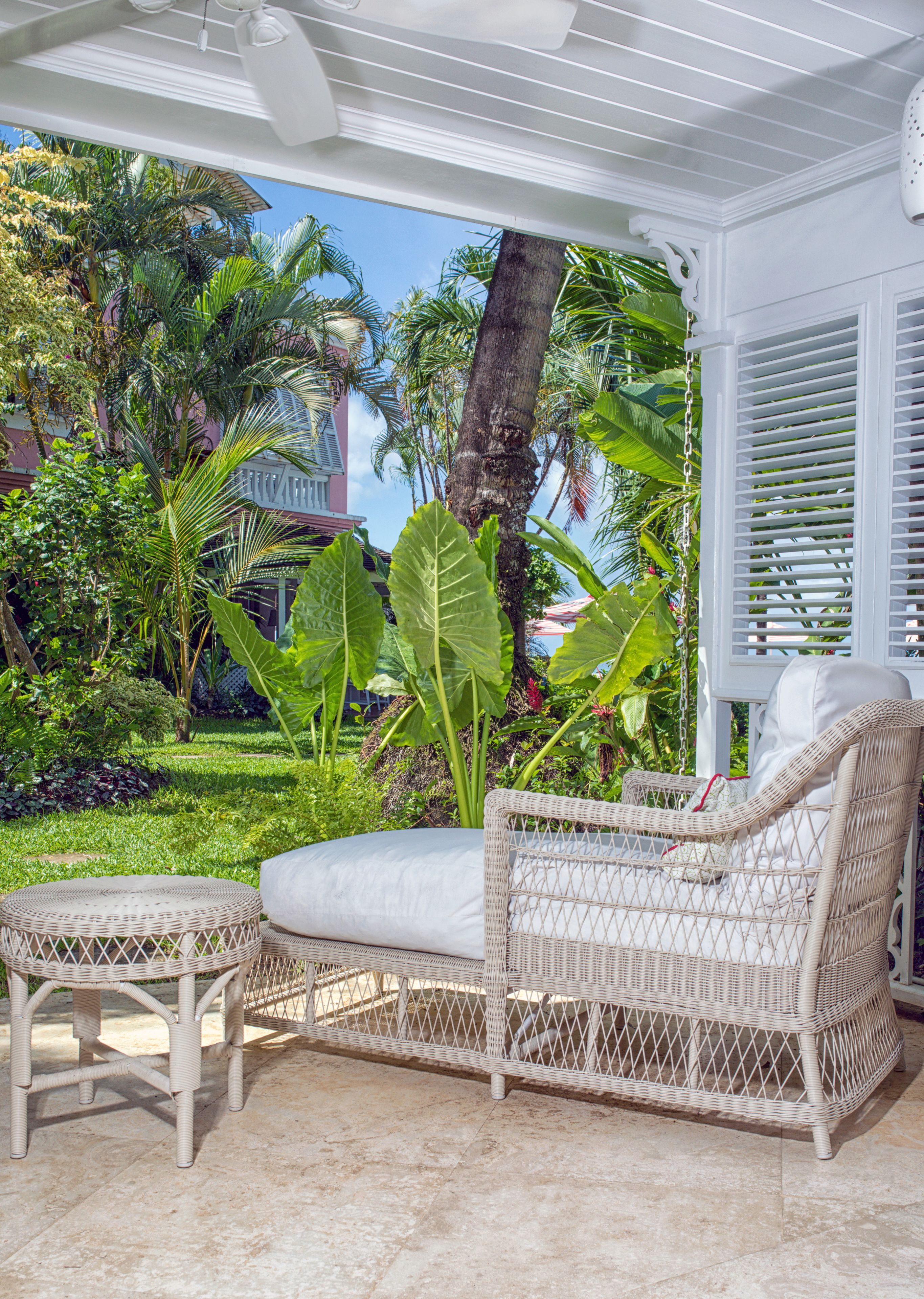Un fauteuil de jardin en osier avec un coussin, un petit pouf rond et un ventilateur de plafond, le tout installé dans un espace extérieur abrité offrant une vue sur des plantes tropicales et un bâtiment en arrière-plan.
