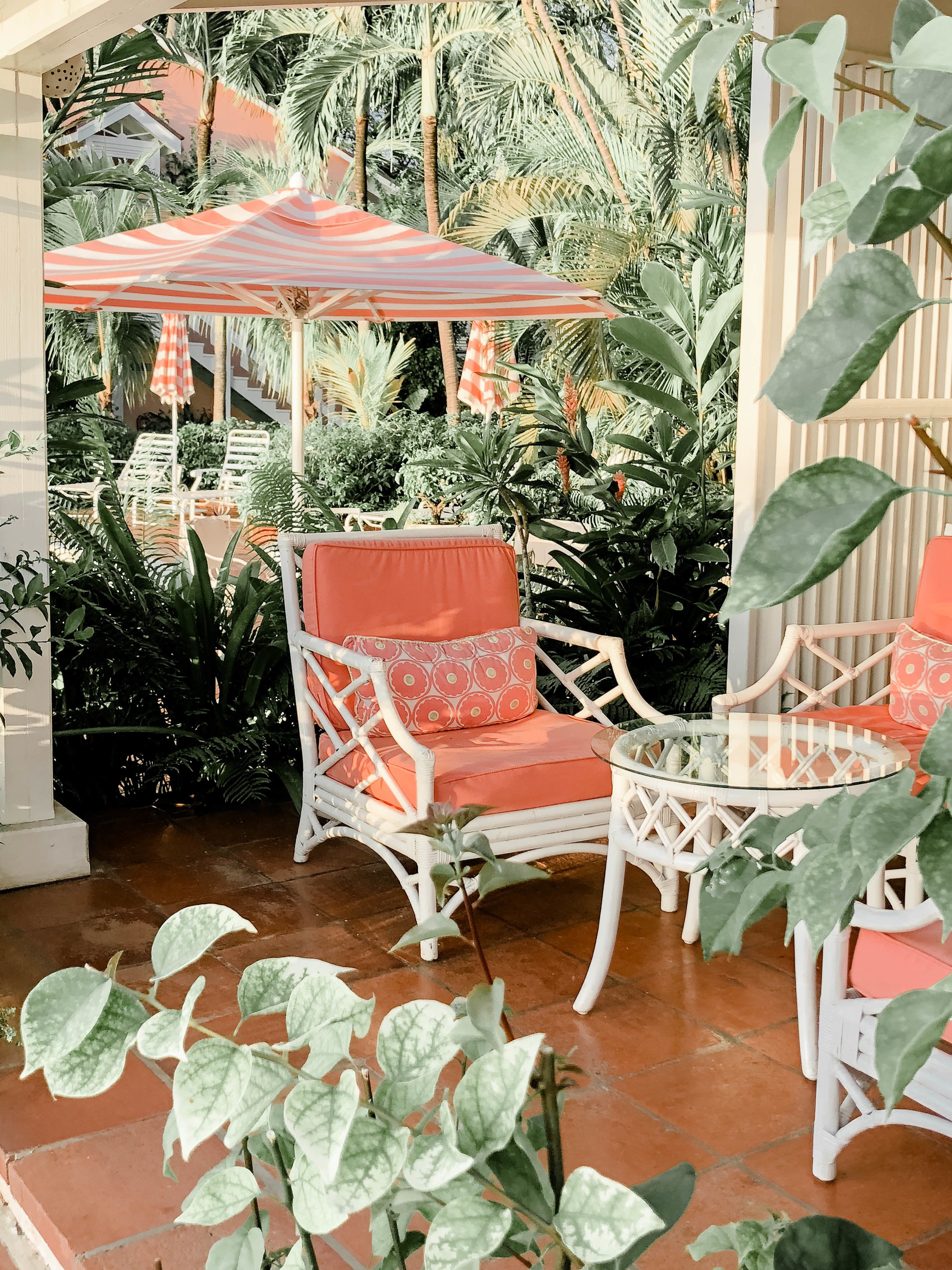 A patio area with wicker chairs and a table, surrounded by lush tropical plants and a striped umbrella.