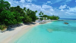 A beach with clear turquoise waters, a sandy shore, and lush green vegetation. There are structures with umbrellas and a building with a red roof. - Cobblers Cove - Barbados (Speightstown)