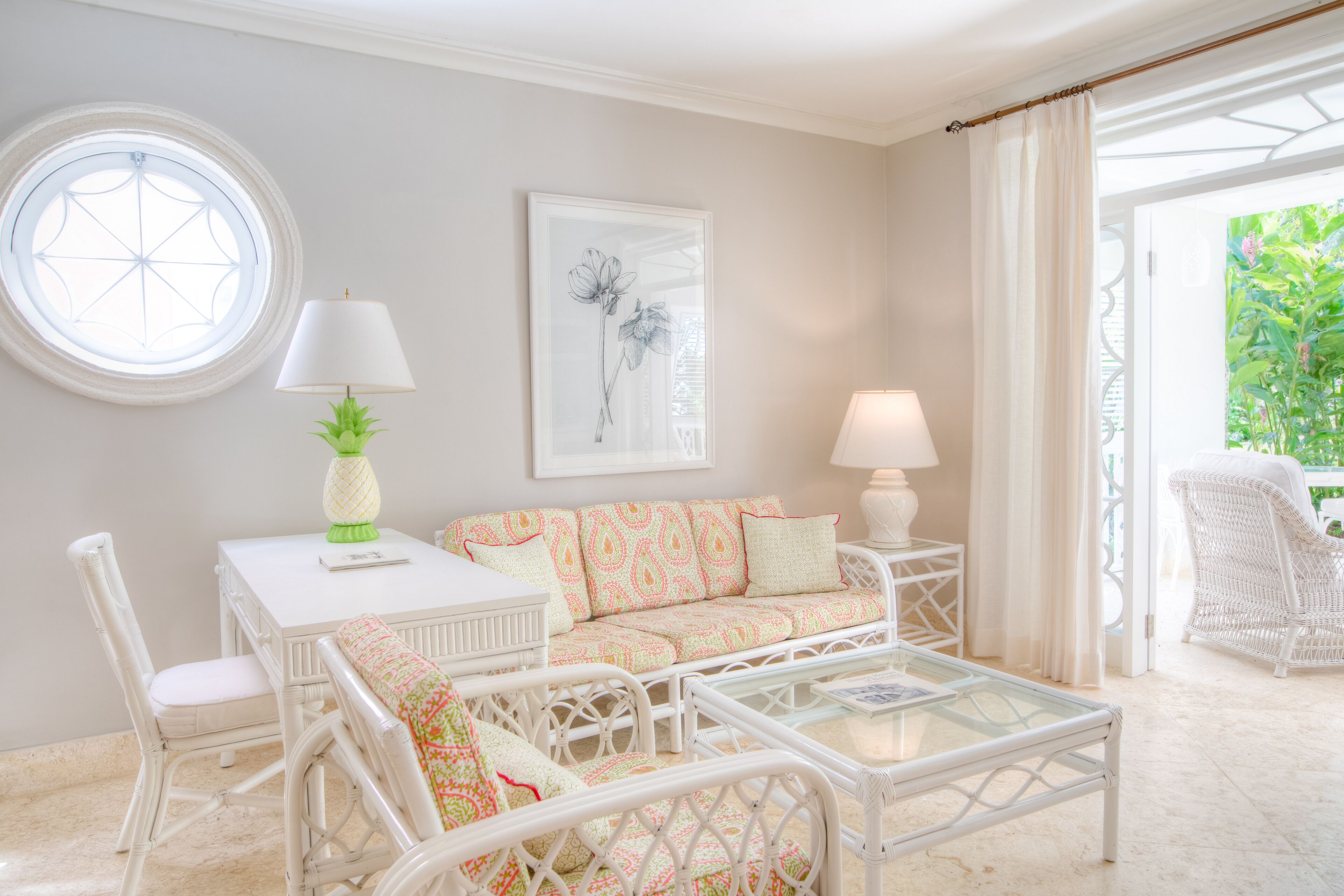 Une pièce avec une fenêtre ronde, un canapé blanc orné de coussins à motifs, une table blanche avec une lampe et un fauteuil en osier.