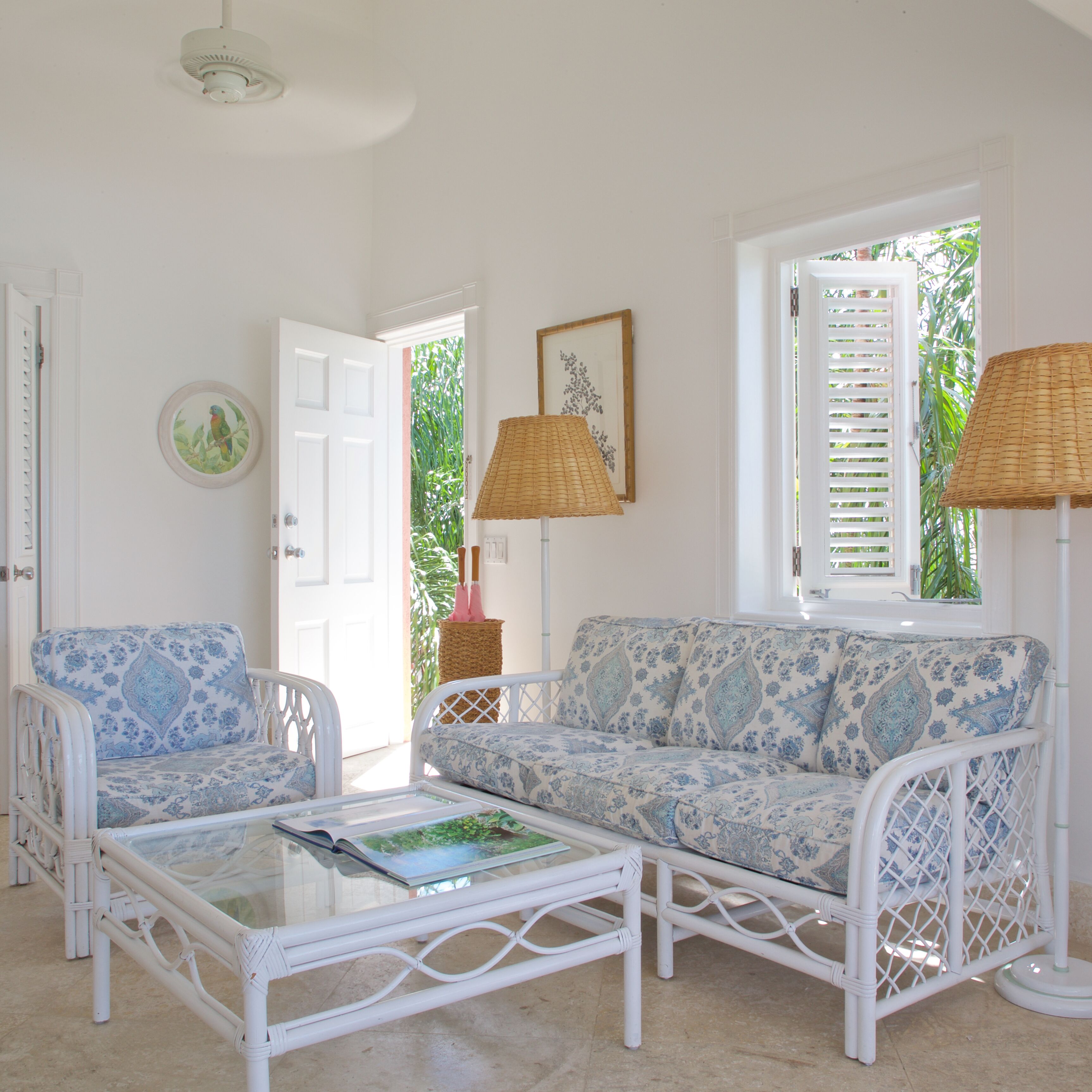 Une pièce avec un canapé blanc, un fauteuil blanc et une table basse en verre. Une porte donne accès à un espace extérieur agrémenté de plantes.