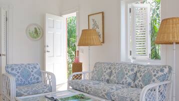 Une pièce avec un canapé blanc, un fauteuil blanc et une table basse en verre. Une porte donne accès à un espace extérieur agrémenté de plantes.