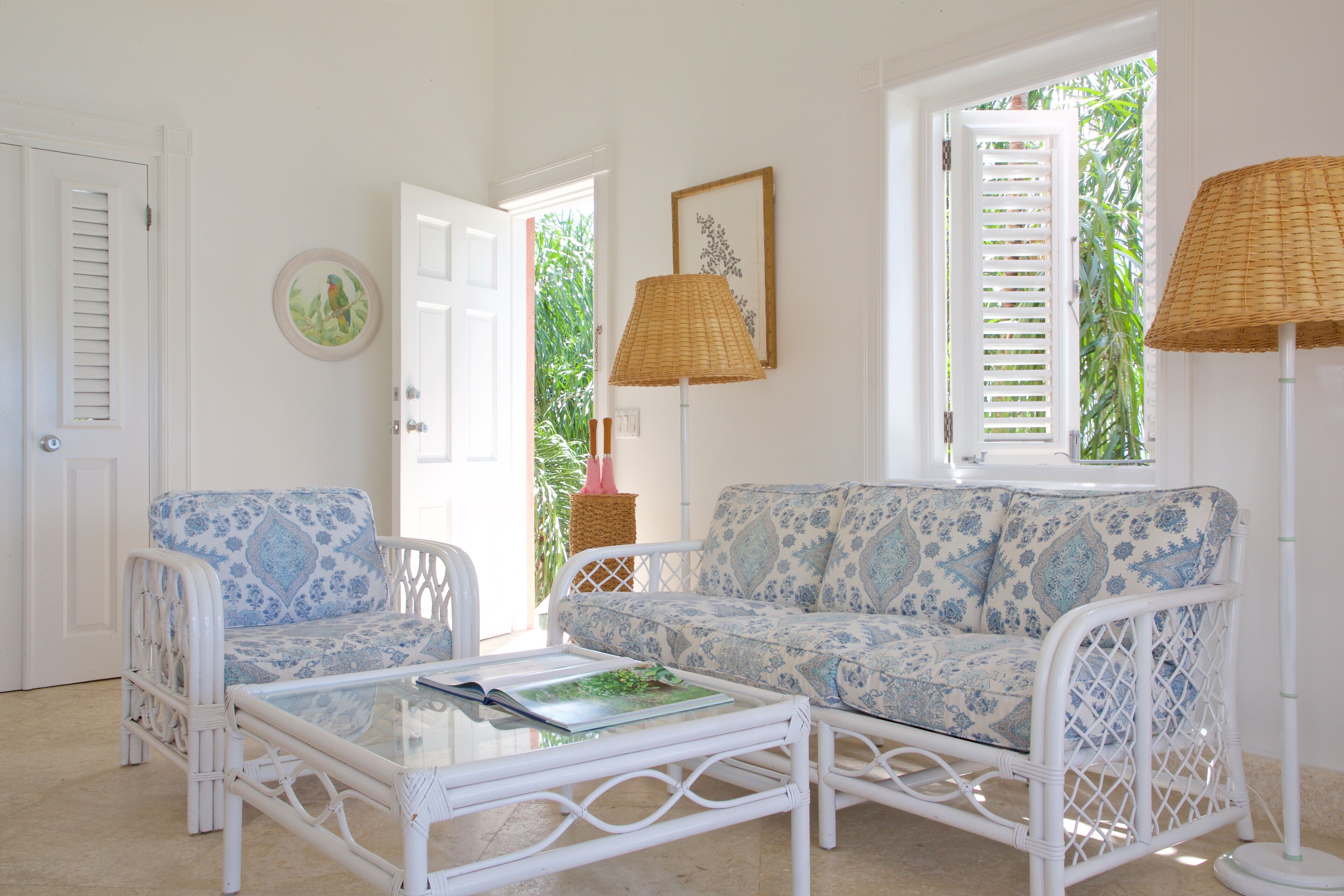 A room with a white wicker sofa, a matching armchair, and a glass coffee table. There is a wicker floor lamp and a framed picture on the wall.