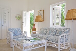 A room with a white wicker sofa, a matching armchair, and a glass coffee table. There is a wicker floor lamp and a framed picture on the wall.