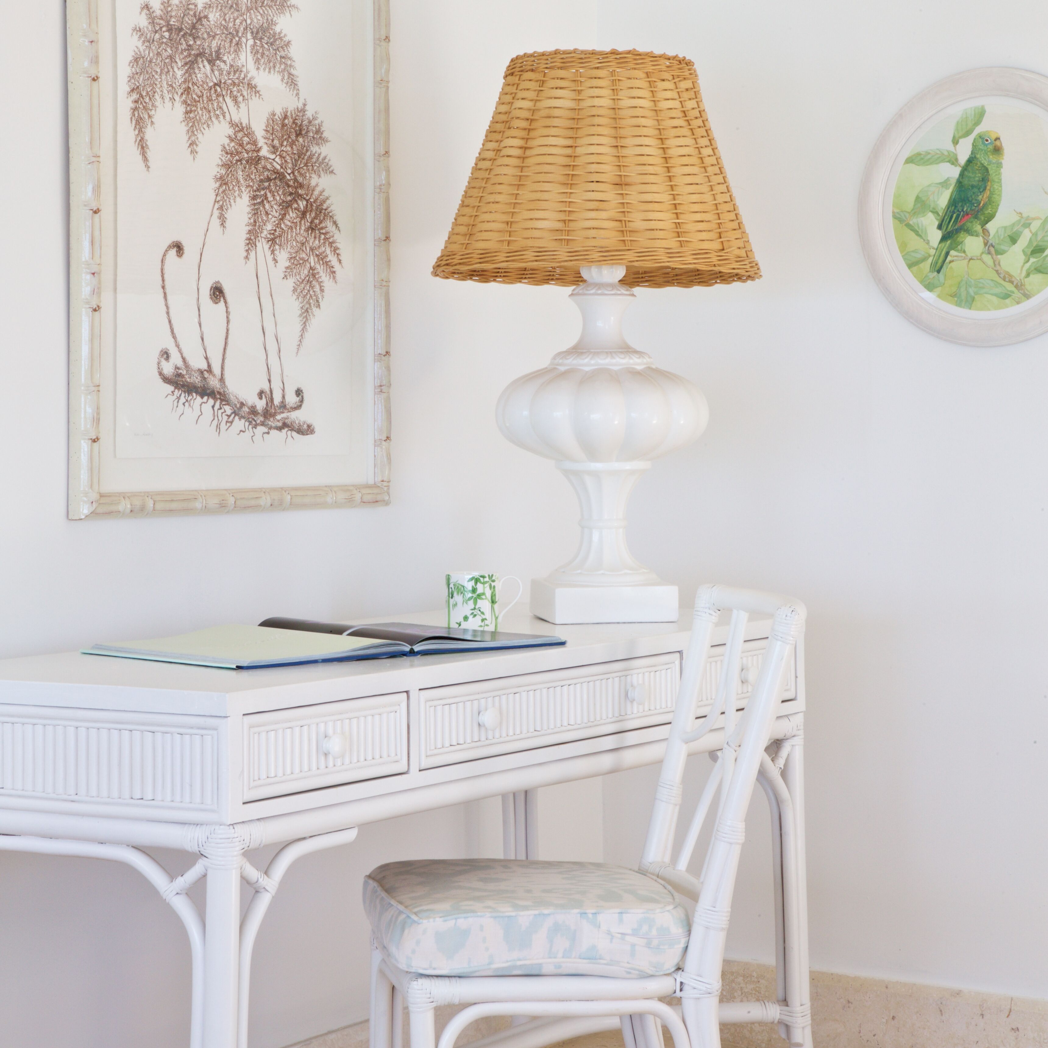 A white desk with a wicker lamp, a framed botanical print, and a small plant.