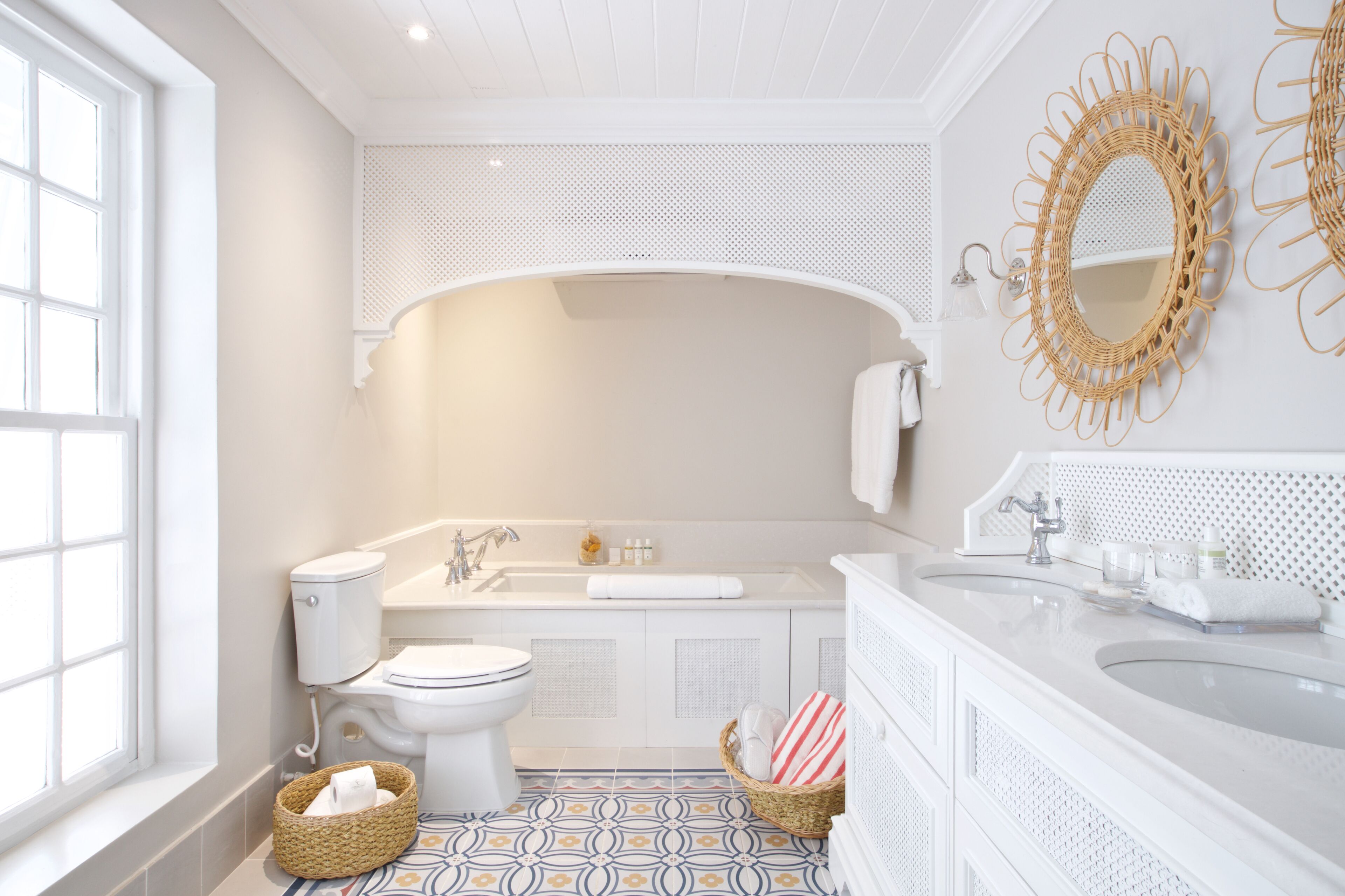 A bathroom with a toilet, sink, and bathtub. There is a window on the left, a decorative mirror on the right, and a patterned floor.