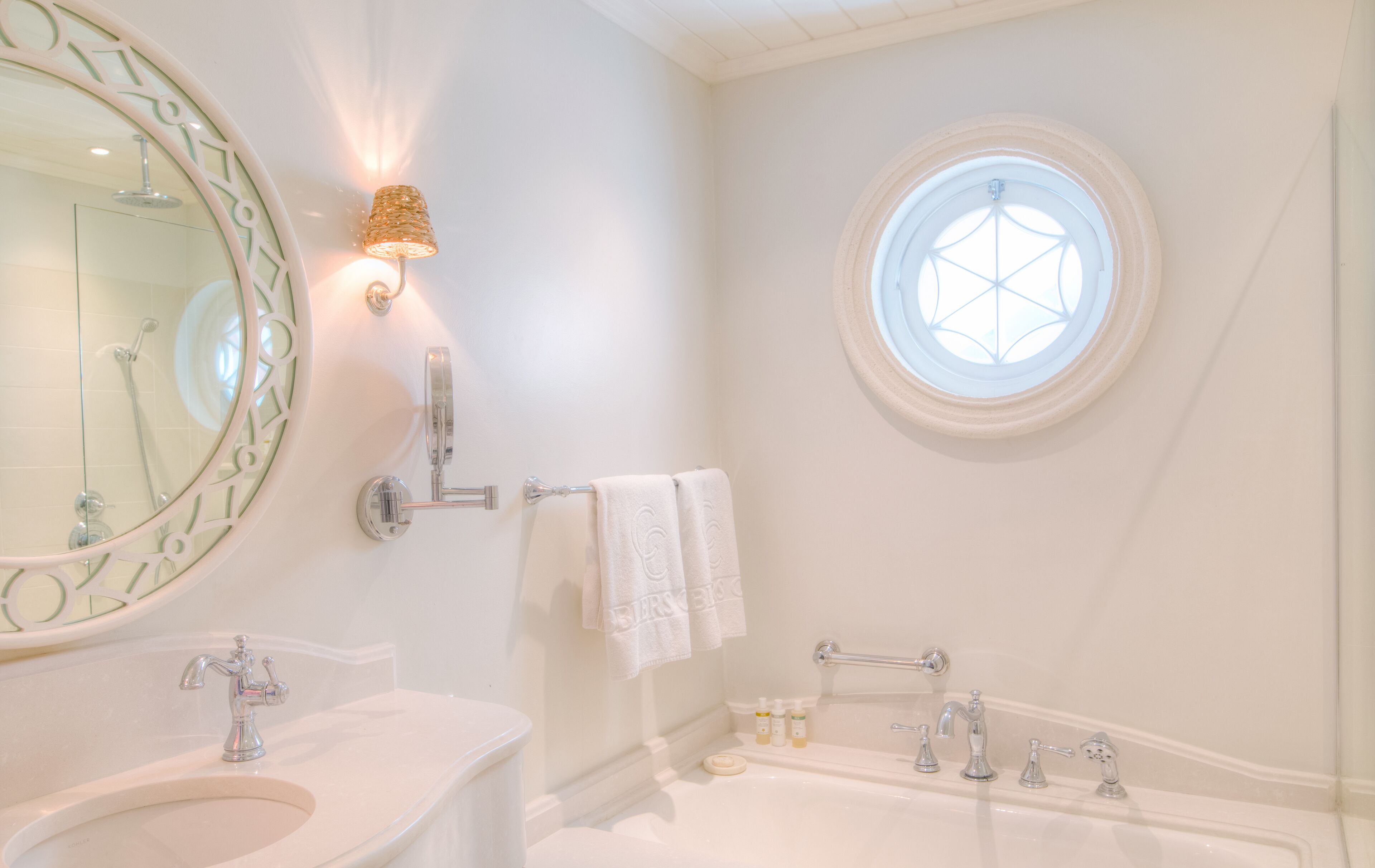 Un baño con un espejo redondo, una lámpara en la pared, un ventanal circular, una bañera y un lavabo.