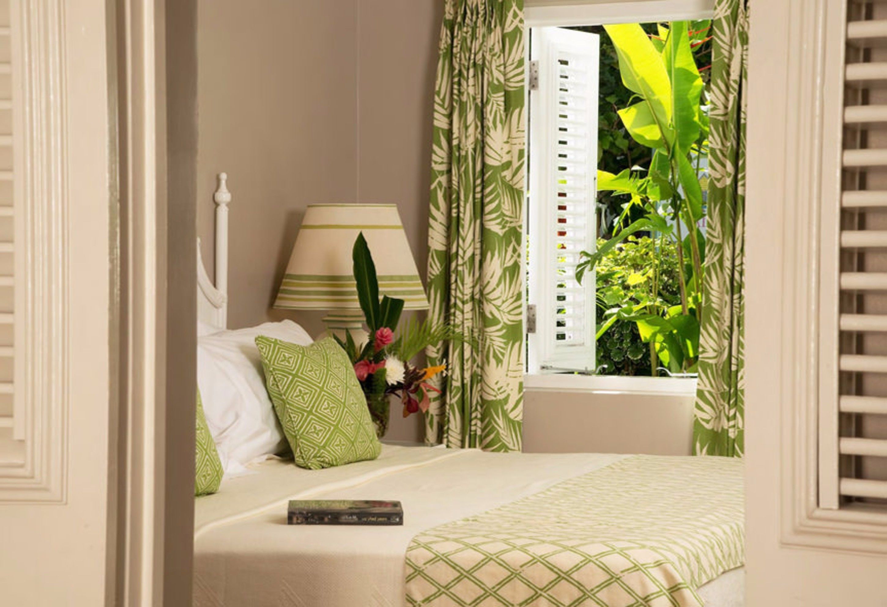 A bedroom with a bed, a nightstand, a lamp, a window with curtains, and a view of greenery outside.