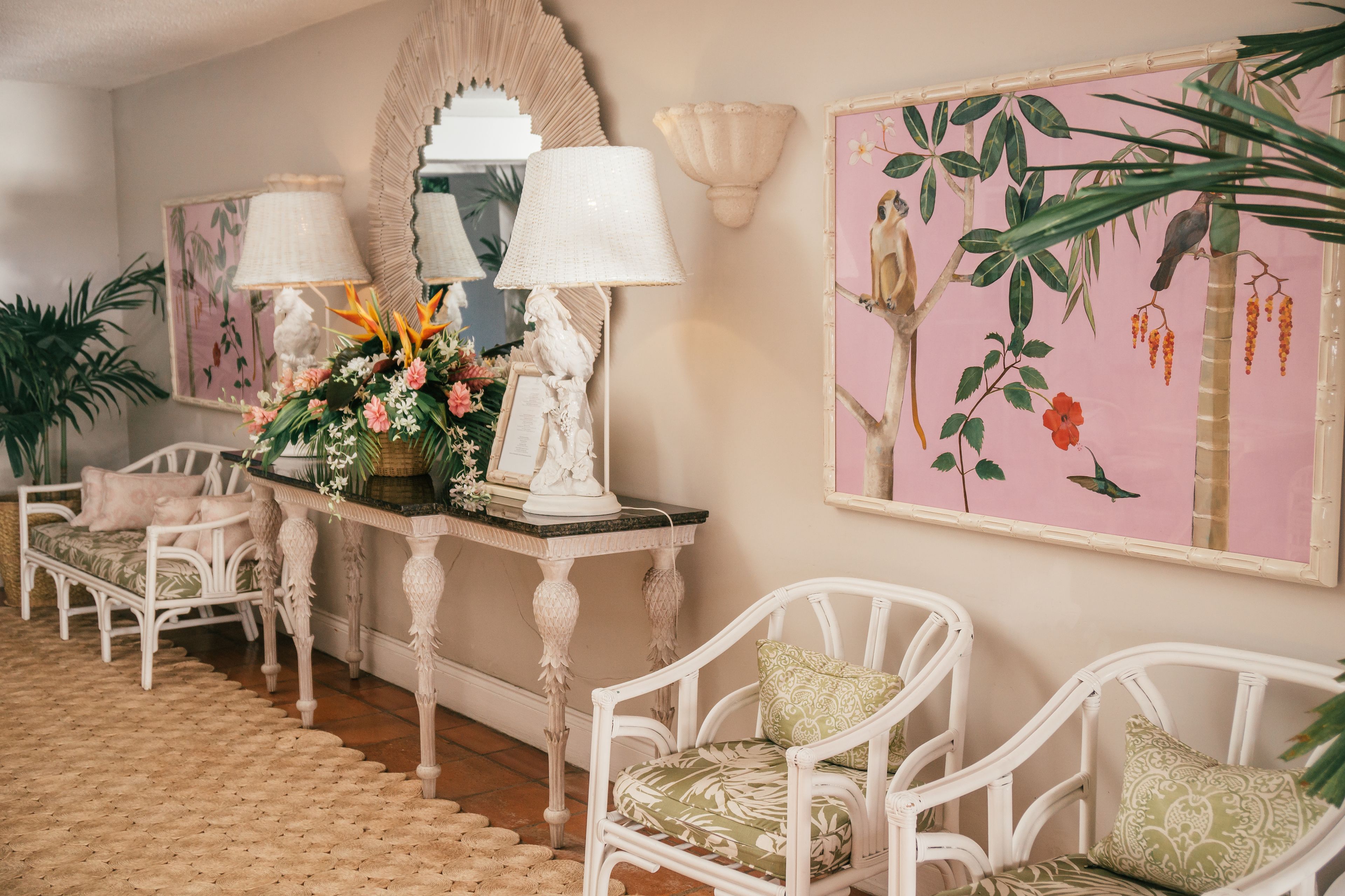 Una habitación con un sofá blanco de mimbre, una mesa con una lámpara y una pared decorada con un cuadro floral.