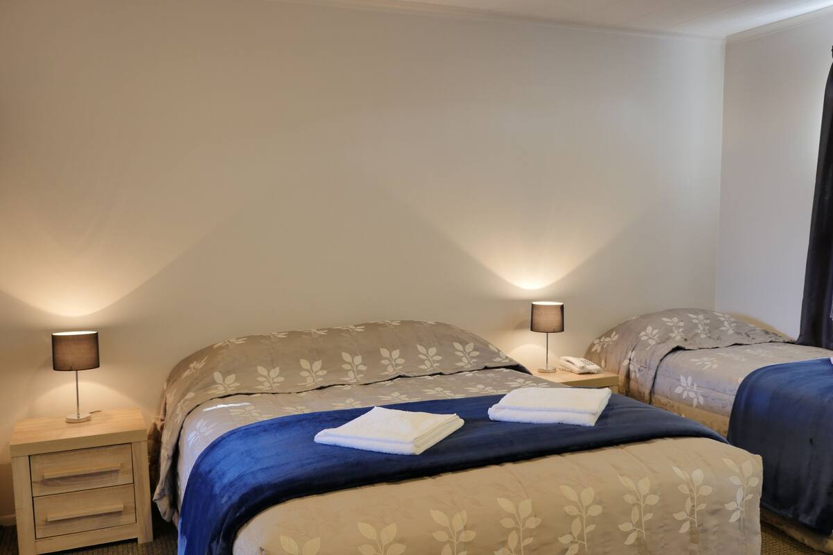Ein Hotelzimmer mit zwei Betten, jeweils mit einer blau-beigen Blumenbettdecke, einem Nachttisch mit Lampe und einer kleinen Schublade.
