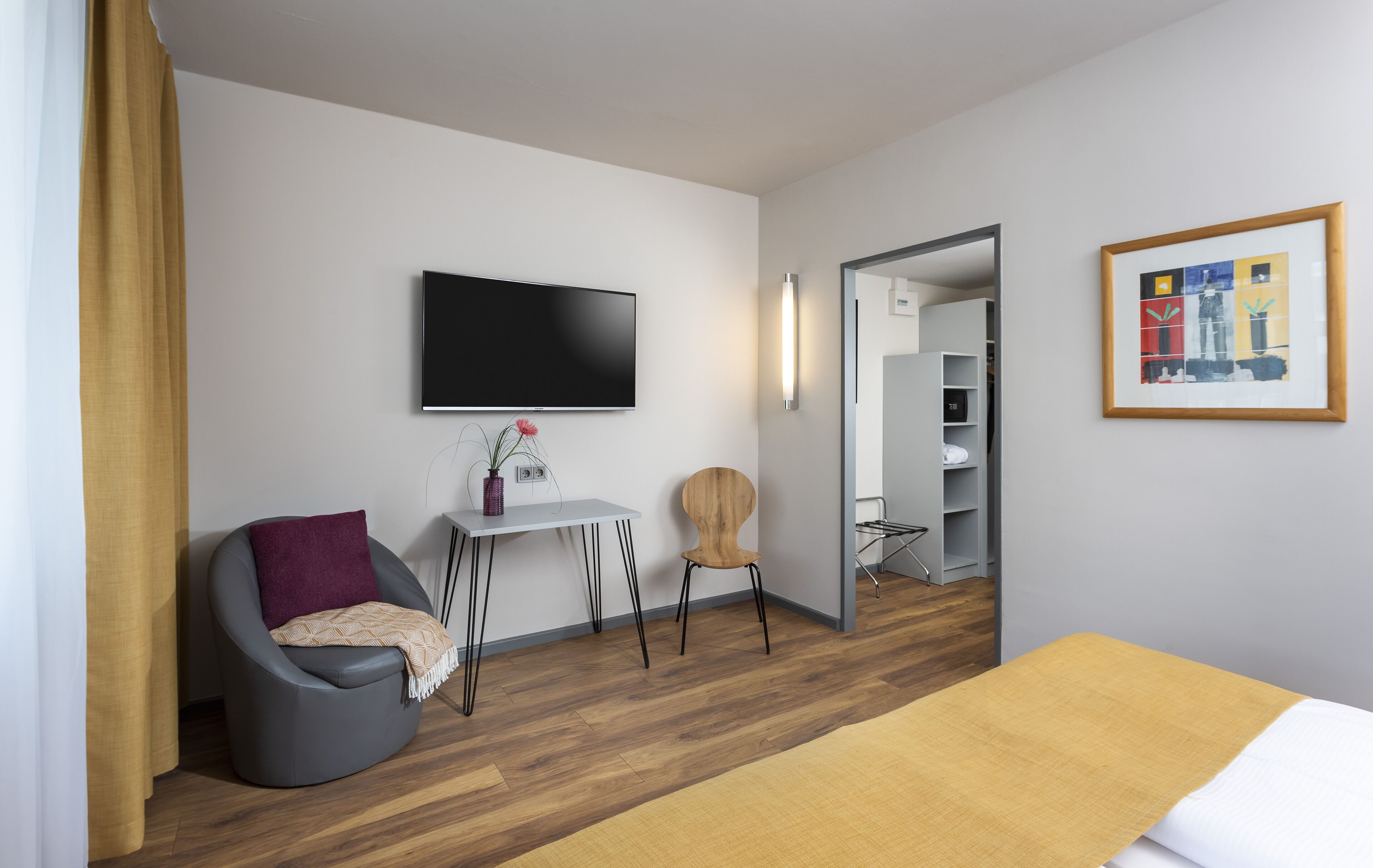 Et moderne hotelværelse med et fladskærms-tv ophængt på væggen, et lille skrivebord med stol, en træstol, en seng med et gult plaide og et billede i ramme på væggen.