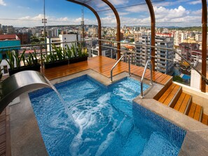 Une piscine sur le toit offrant une vue sur la ville, entourée de plantes et d’une terrasse en bois.