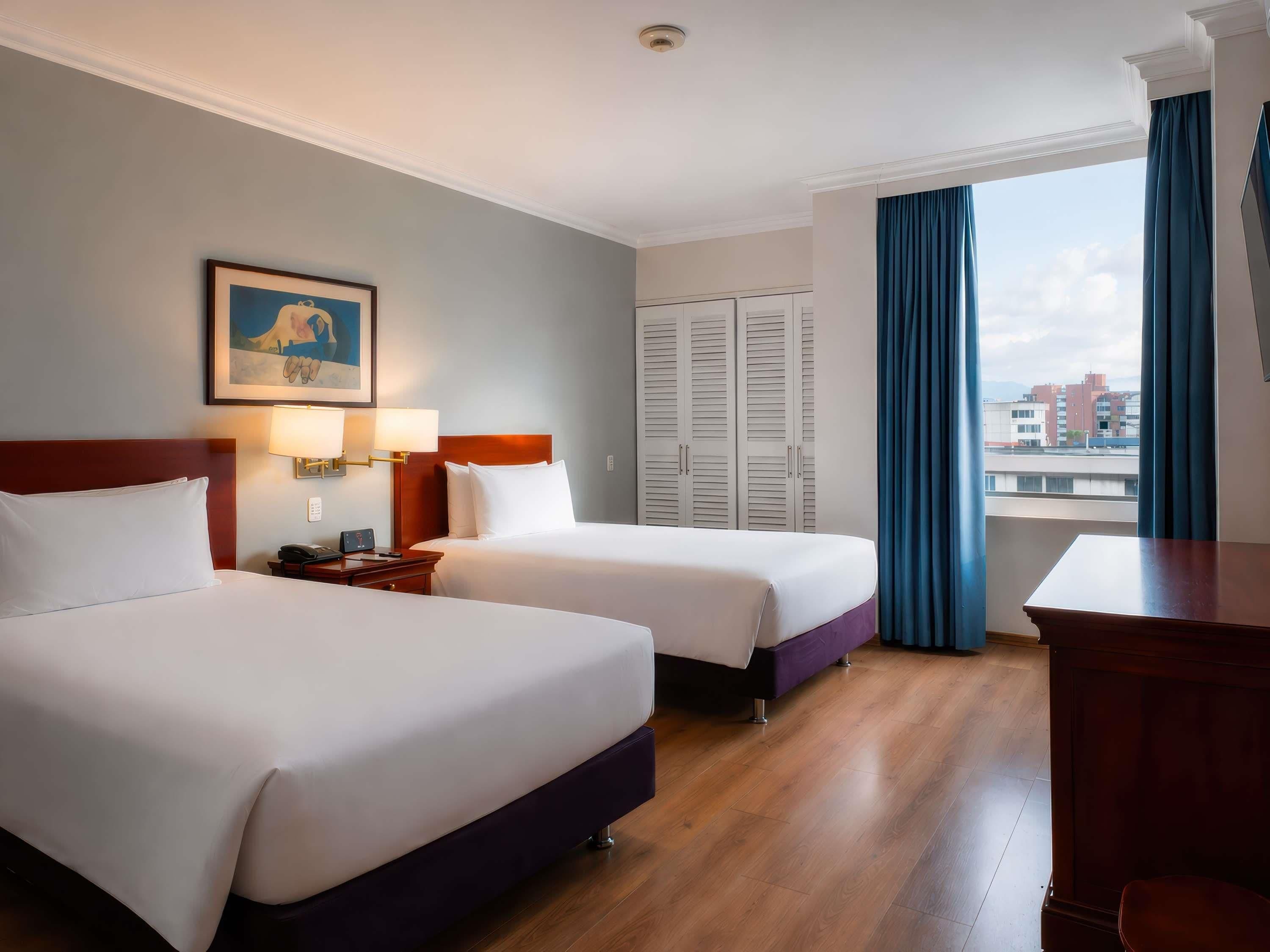 A hotel room with two beds, a desk, a chair, a window with curtains, and a city view.