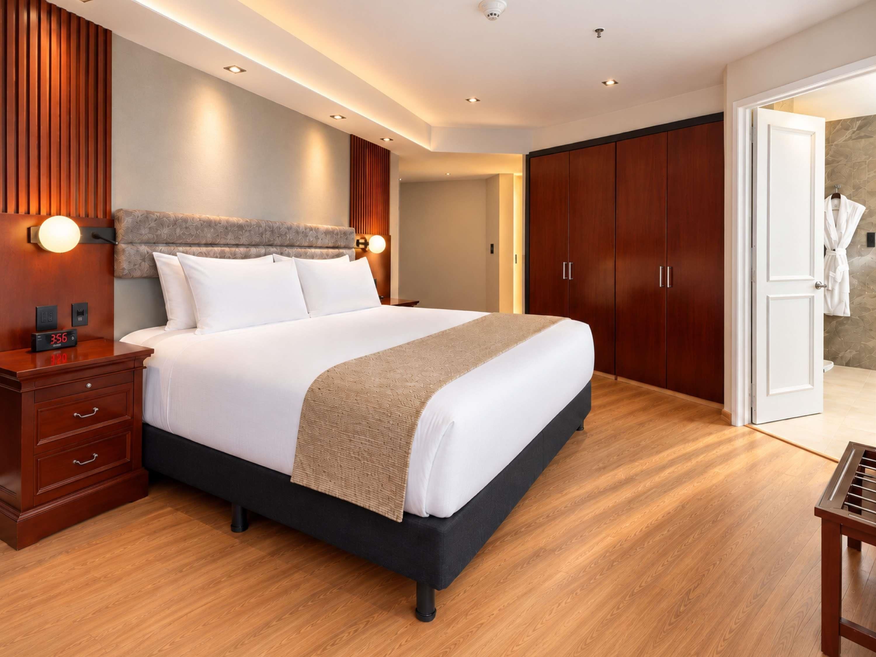 Una habitación de hotel moderna con una cama grande, un armario de madera y un baño visible a través de una puerta abierta.
