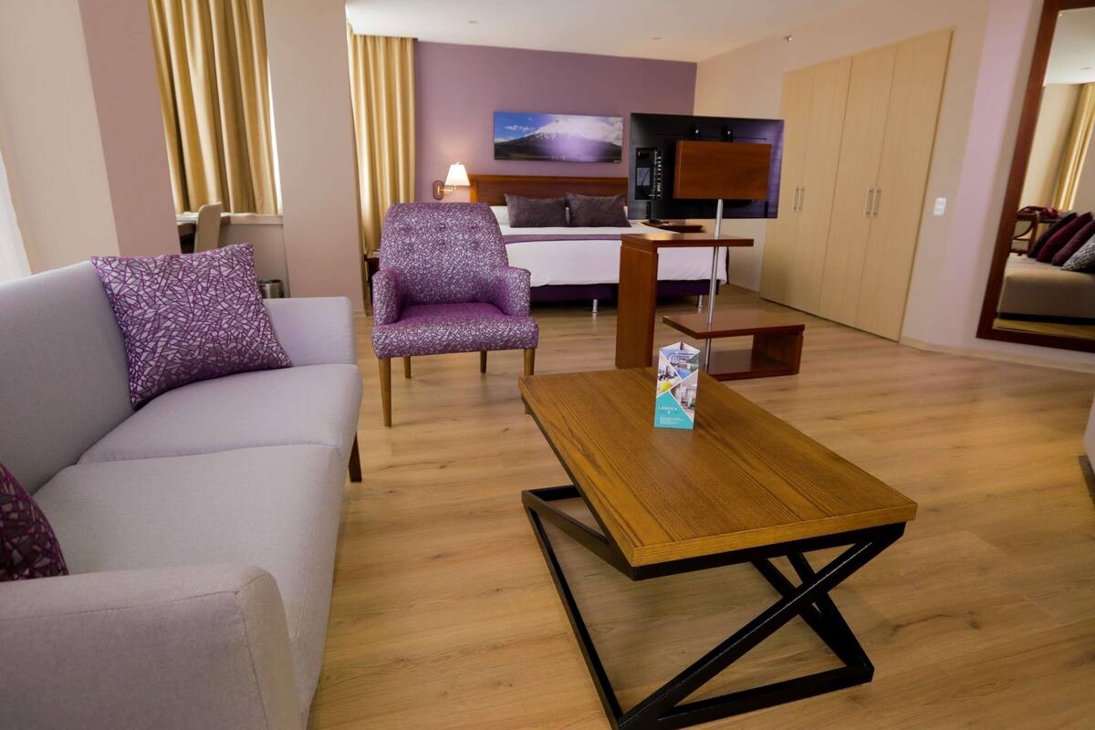 A modern hotel room with a sofa, armchair, wooden coffee table, TV, and a kitchen area in the background.