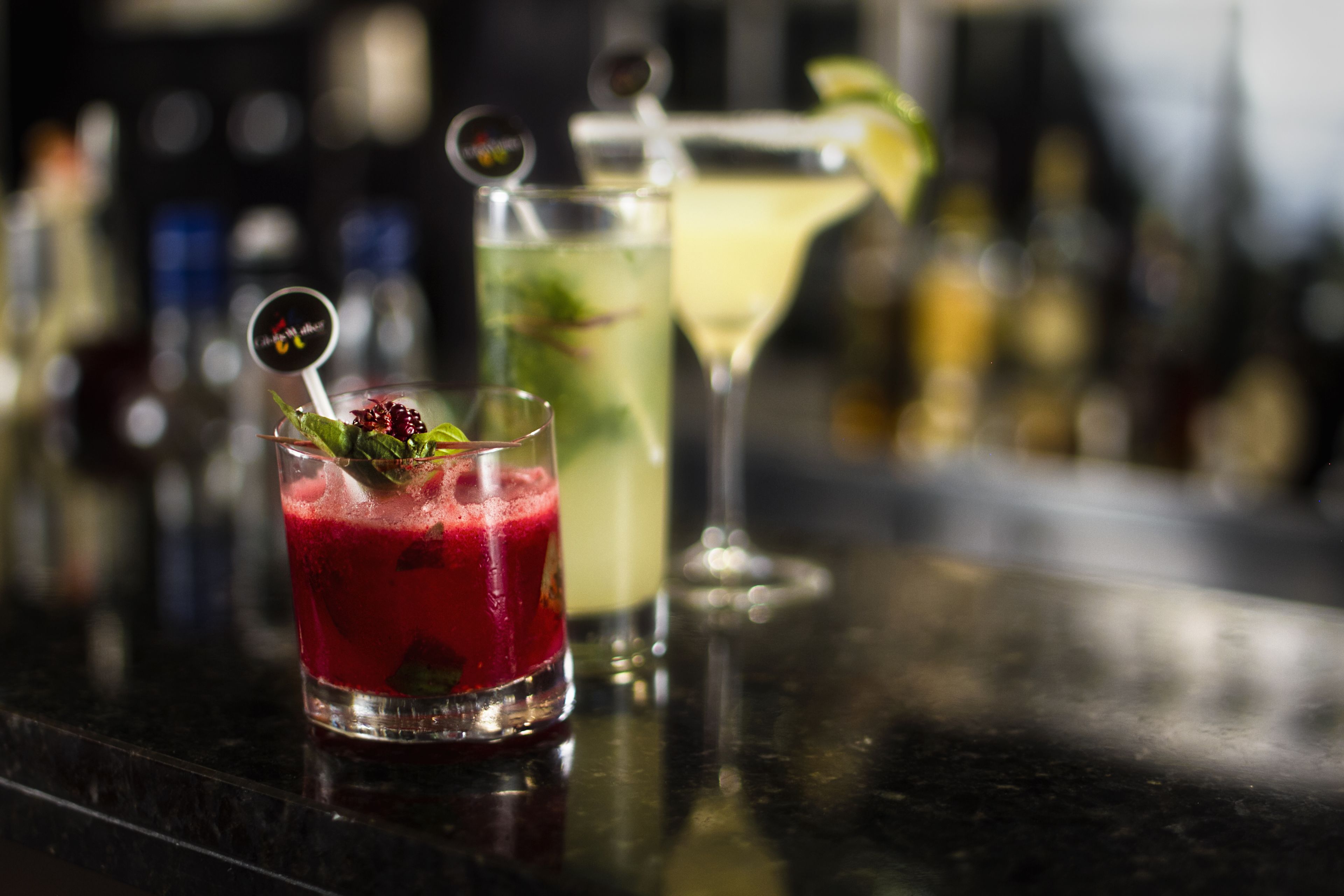 Three cocktails on a bar counter with a blurred background of bottles.