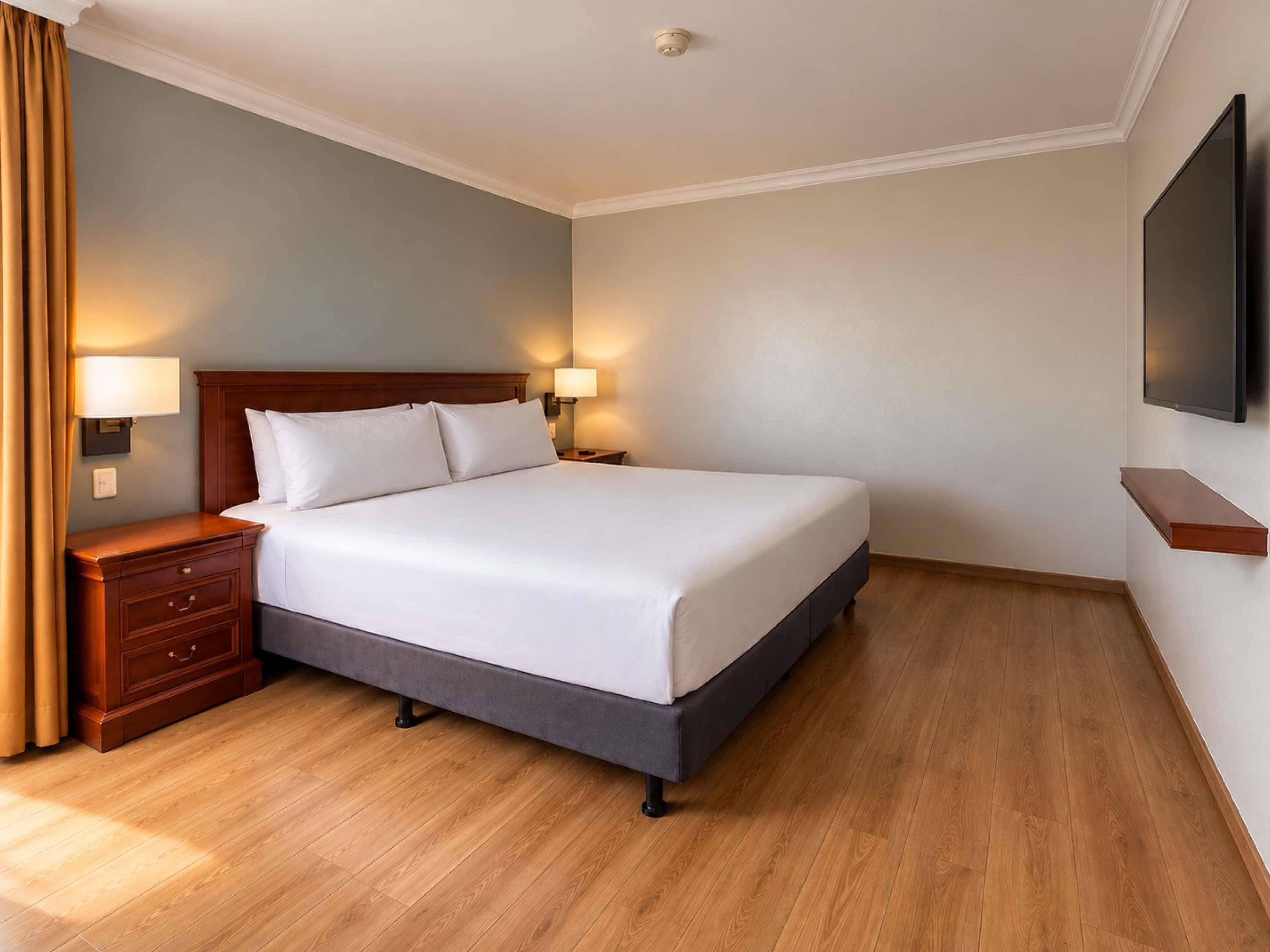 A hotel room with a bed, bedside tables, lamps, a flat-screen TV, and wooden flooring.
