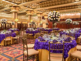 Un salón de banquetes con mesas redondas cubiertas de manteles morados, sillas doradas y centros de mesa florales.
