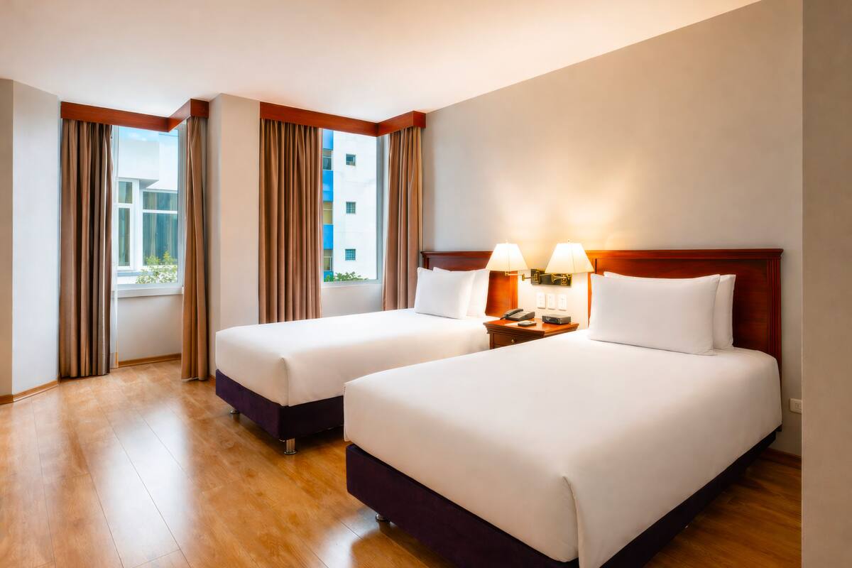 A hotel room with two beds, a wooden floor, a window with curtains, and a nightstand with lamps.