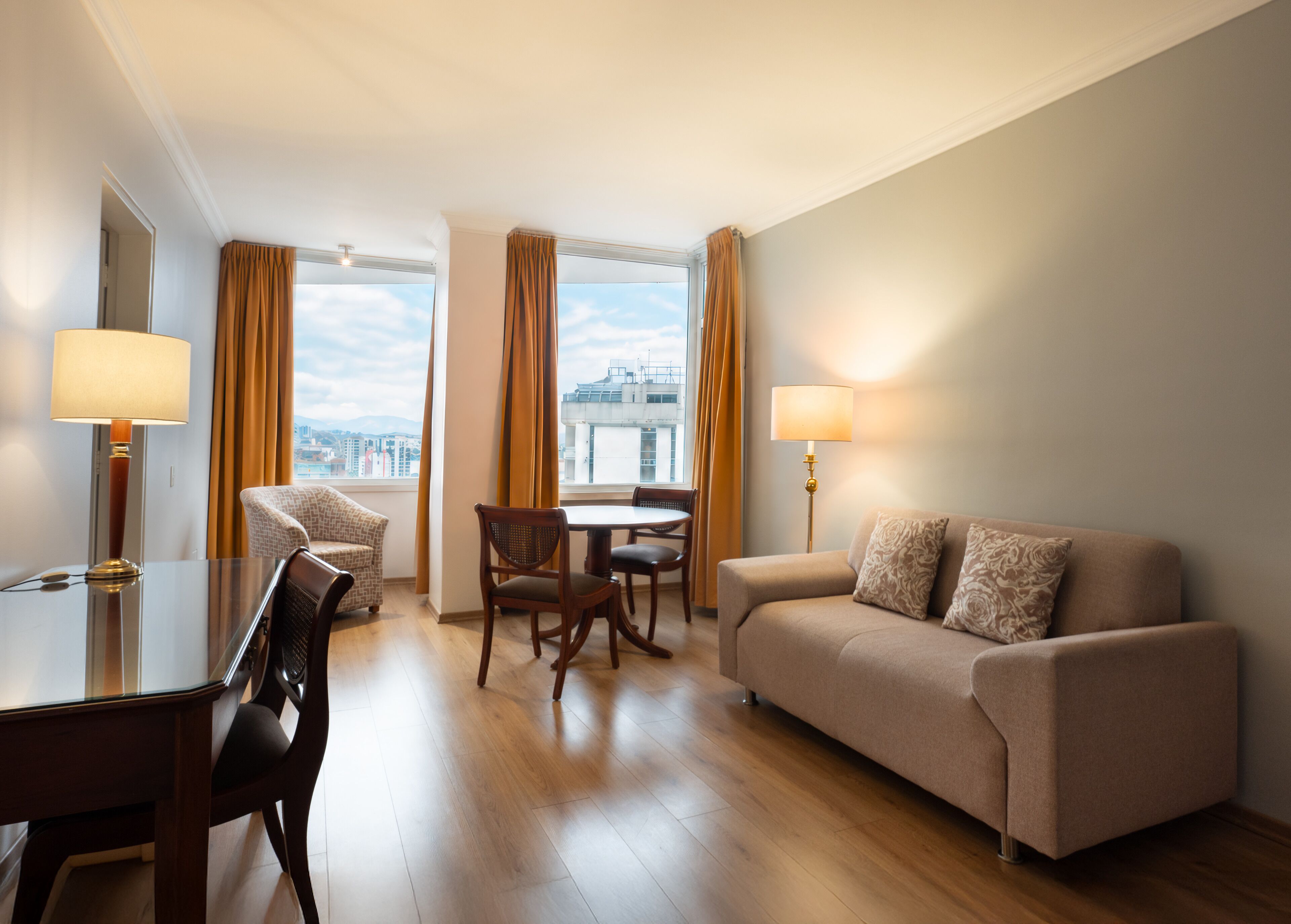 A hotel room with a sofa, a dining table with chairs, a lamp, and large windows with curtains.