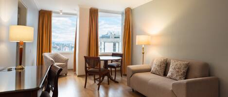 A hotel room with a sofa, a dining table with chairs, a lamp, and large windows with curtains.