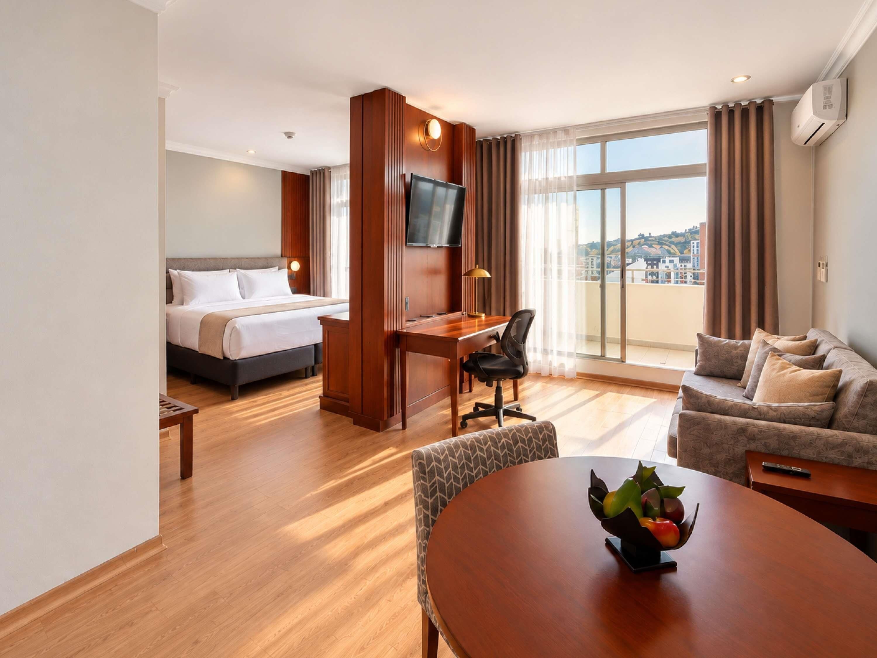 Une chambre d’hôtel équipée d’un lit, d’un bureau, d’une télévision, d’un canapé et d’une table à manger avec des fruits.