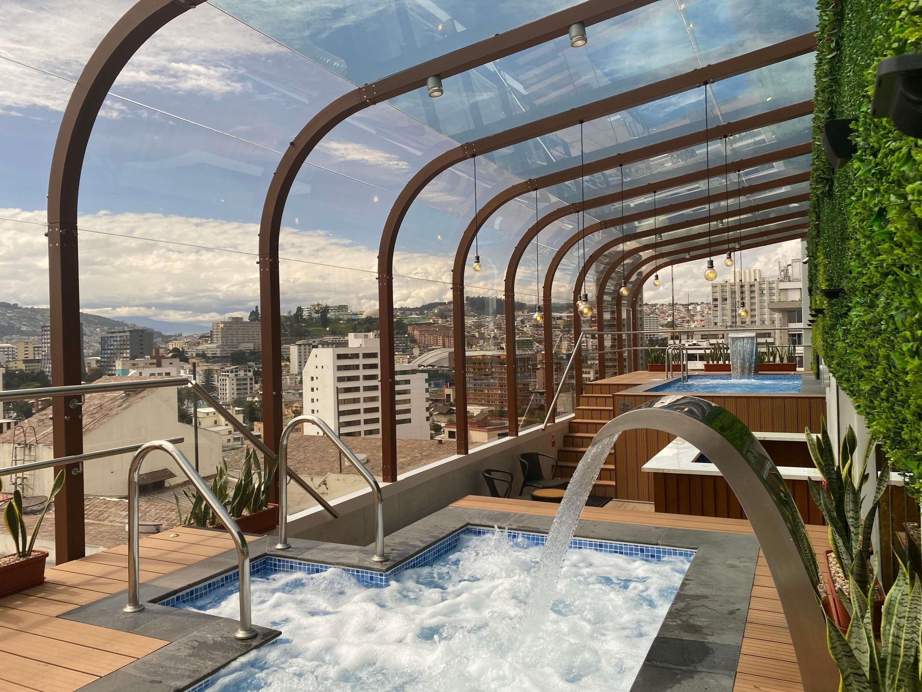 A rooftop pool area with a clear view of the cityscape, surrounded by glass railings and potted plants.