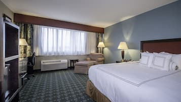 A hotel room with a large bed, a desk with a chair, a television, a window with curtains, and two bedside lamps.