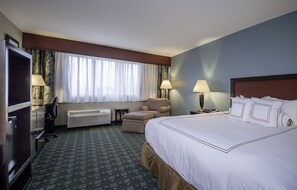 A hotel room with a large bed, a desk with a chair, a television, a window with curtains, and two bedside lamps. - Causeway Bay Hotel (Lansing)