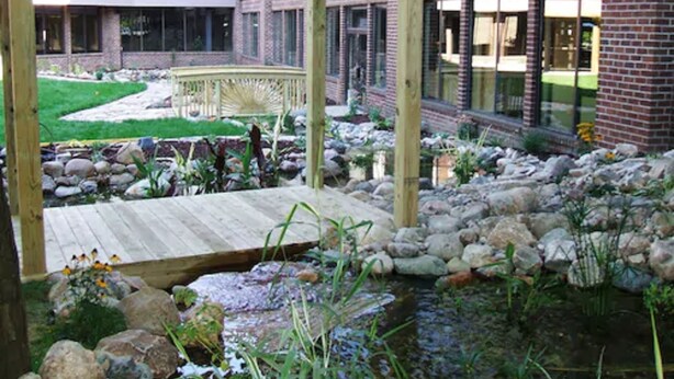 A wooden deck leading to a garden area with a pond, surrounded by a brick building with large windows. - Causeway Bay Hotel (Lansing)