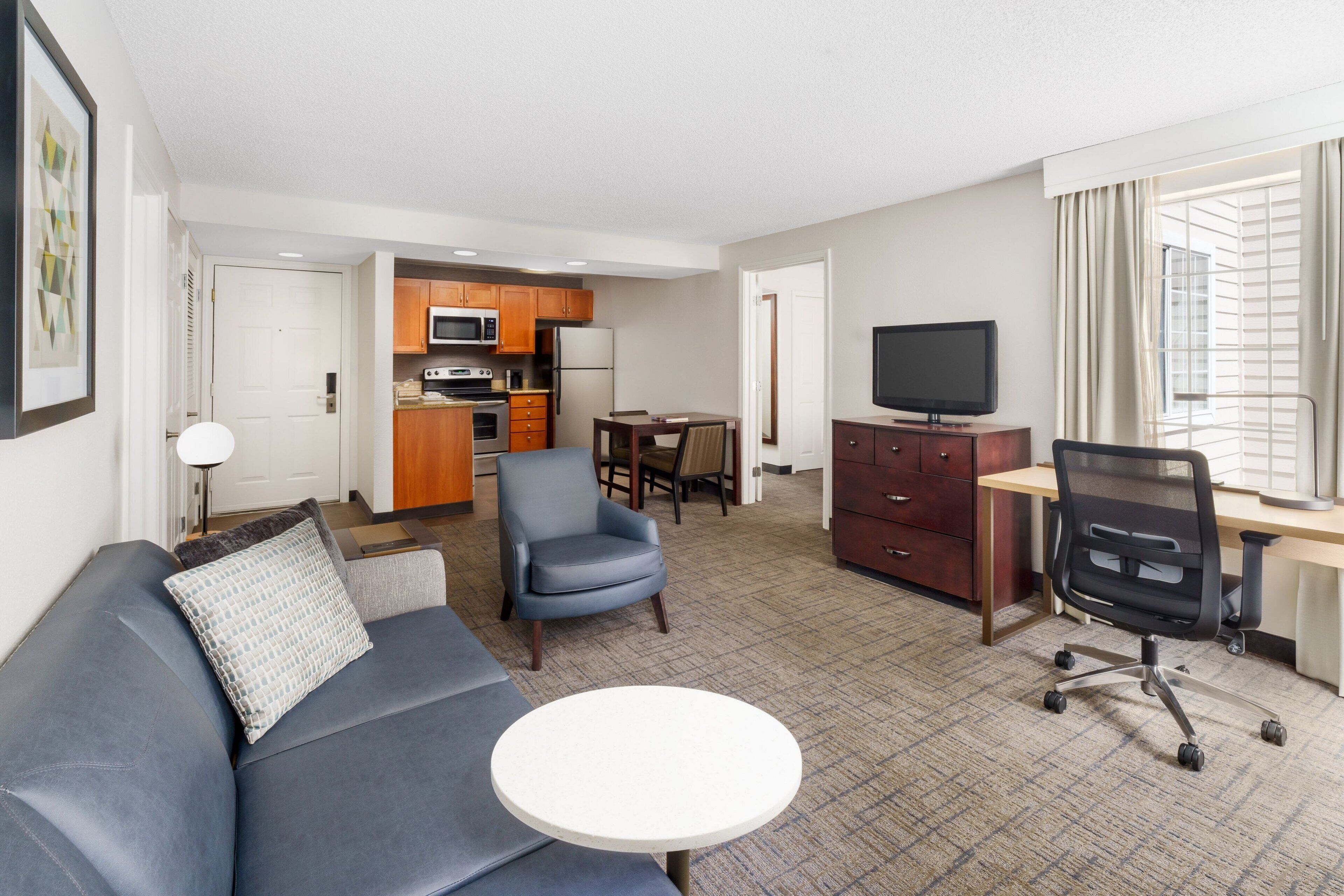A hotel room with a sofa, a round table, a TV, a desk, and a kitchen area.