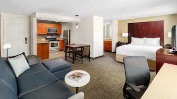 A hotel room with a bed, a sofa, a small dining table, a kitchen area, and a desk.