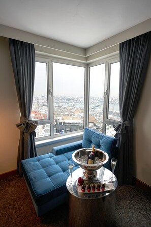 A corner room with a large window, a blue sofa, a small round table with a bottle and glasses, and a plate of chocolates.
