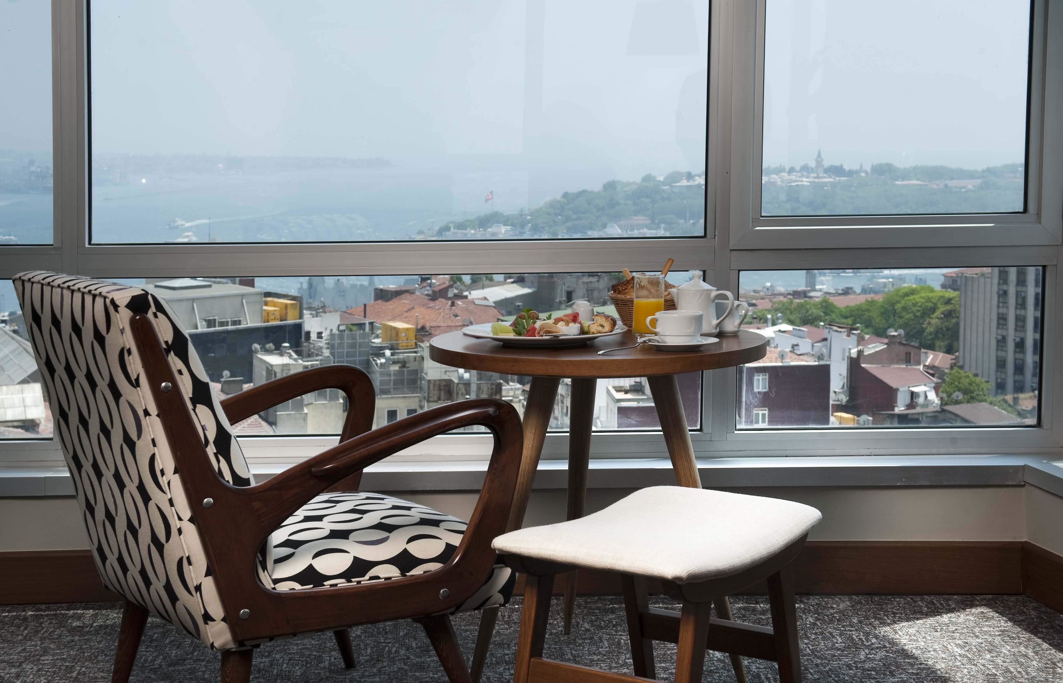 A room with a large window, a wooden chair with a patterned cushion, a small round table with breakfast items, and a view of a cityscape and water.