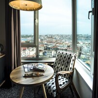 Una habitaci ón con un ventanal grande que ofrece vistas a la ciudad, una mesa redonda de madera con una revista y una silla estampada.
