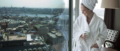 Una persona con una bata blanca y una toalla en la cabeza, sosteniendo una taza, de pie junto a una ventana con vista a un paisaje urbano.
