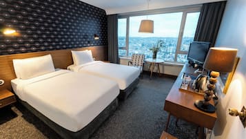 Ein Hotelzimmer mit zwei Betten, einem Schreibtisch mit Fernseher, einem Fenster mit Blick auf die Stadt und einer Lampe.