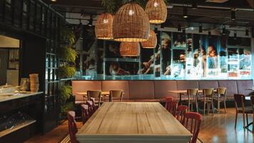 A restaurant with a long wooden table, red chairs, patterned floor tiles, hanging wicker lamps, and a glass partition with a view of the kitchen.