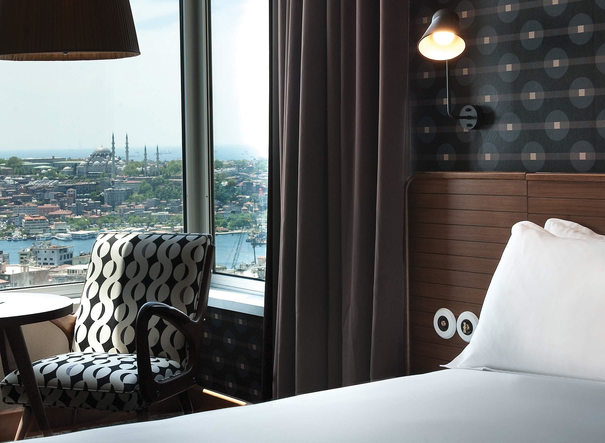 A hotel room with a large window offering a city view, a patterned armchair, a bedside table, and a lamp.