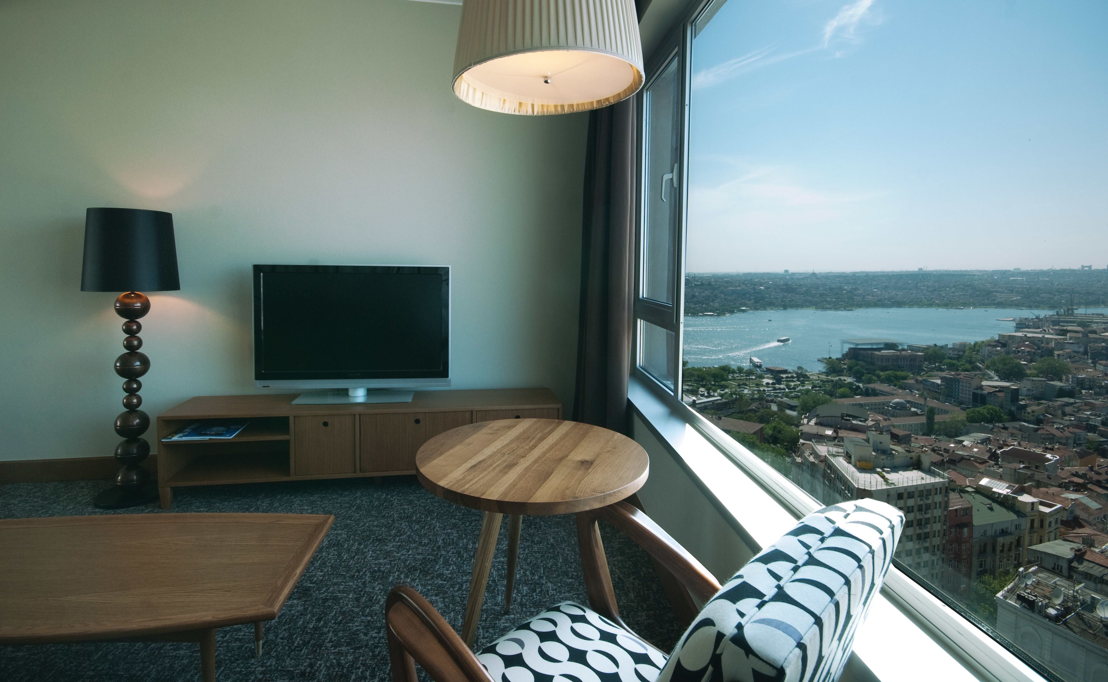 A modern living room with a TV, wooden furniture, and a large window offering a city view.