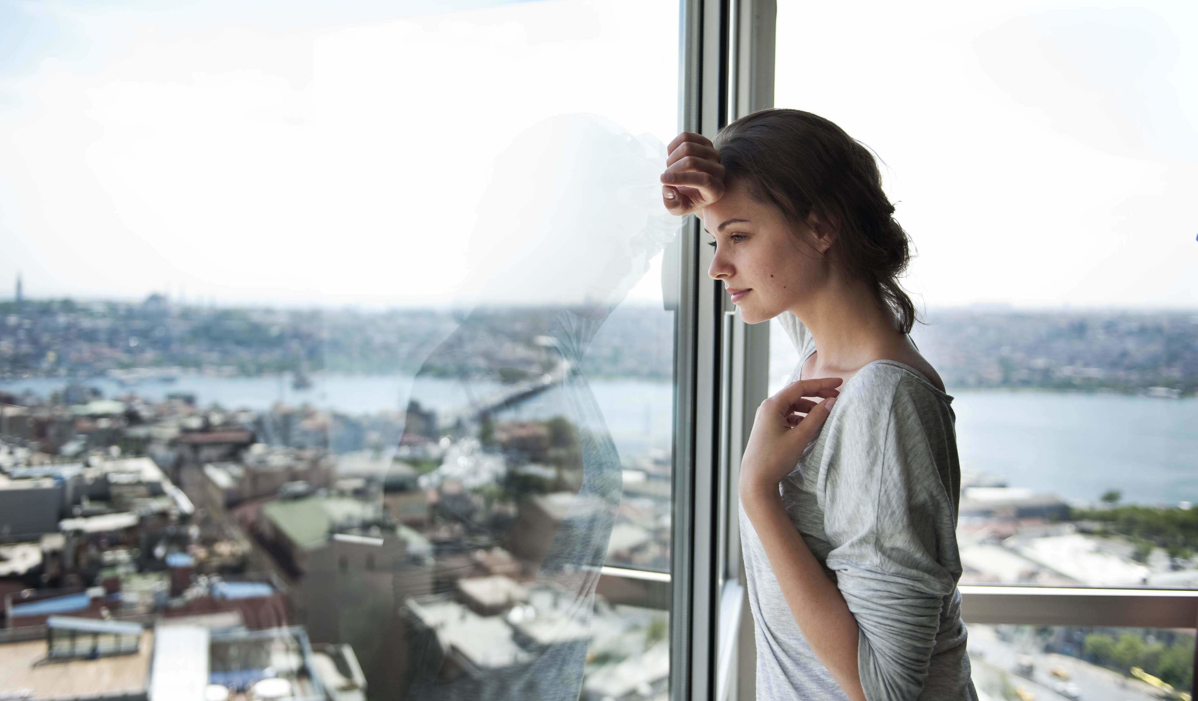 A person standing by a window overlooking a cityscape with buildings and a body of water.