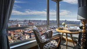 Una habitación con un ventanal que ofrece vista a la ciudad, una mesa redonda con un florero y una silla con un cojín estampado.