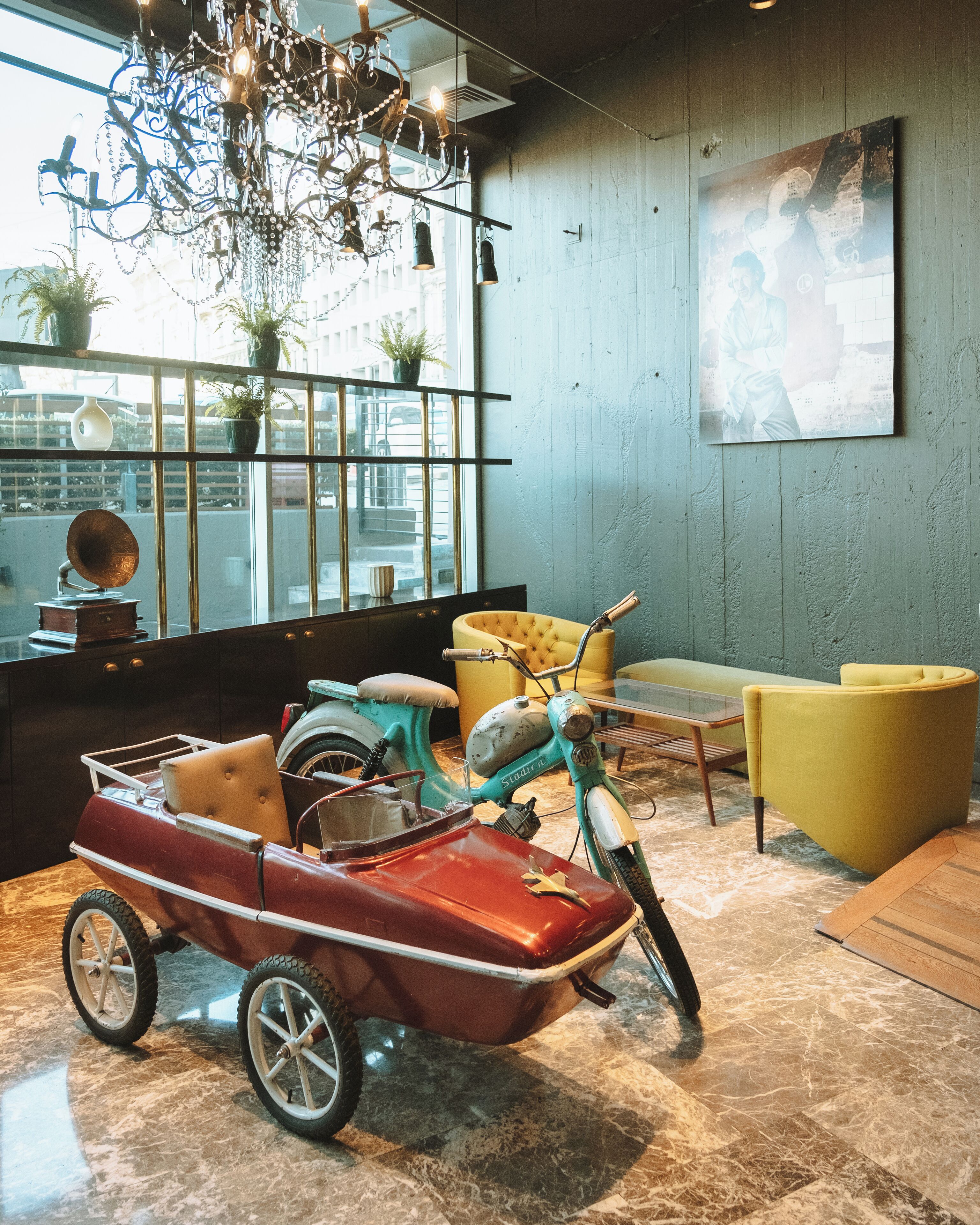 Une voiture ancienne avec un side-car, un fauteuil jaune, un canapé bleu, un lustre et une grande fenêtre donnant sur la ville.